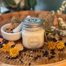 Load image into Gallery viewer, Candle in a jar labeled 'Apothecary' surrounded by flowers and herbs on a woven surface.