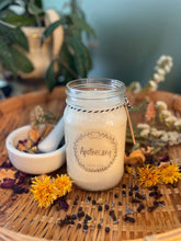 Load image into Gallery viewer, Candle in a glass jar labeled 'Apothecary' on a wooden surface with flowers and herbs.