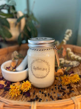 Load image into Gallery viewer, Mason jar candle labeled 'Apothecary' on a woven mat with flowers and herbs