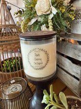 Load image into Gallery viewer, Candle labeled &#39;Dark Cherry Orchard&#39; on a wooden stand with decorative elements in the background