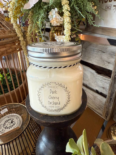 Candle in a mason jar labeled 'Dark Cherry Orchard' on a wooden stand with decorative background